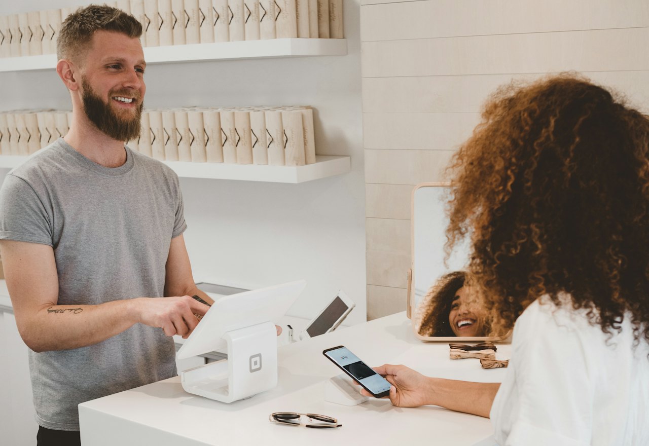 A shop owner at the counter serving a customer who is paying with their phone